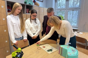 Cztery osoby stoją wokół stołu w jasnej sali lekcyjnej. Dwie uczennice po lewej mają na sobie białe koszule, a obok nich stoi Michał Rajwa Naczelnik wydziału edukacji w bordowej marynarce. Kobieta po prawej pochyla się nad stołem i wskazuje dłonią element leżący na planszy. Na blacie znajdują się materiały dydaktyczne, w tym plansza z rysunkami oraz model w formie kolorowej makiety.