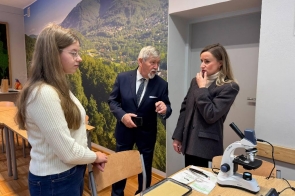 Trzy osoby stoją obok siebie w sali z dużą fototapetą przedstawiającą górski krajobraz. Po lewej znajduje się młoda dziewczyna w jasnym swetrze, pośrodku Stanisław Kubicius Przewodniczący Rady w garniturze wykonuje gest ręką, a po prawej stoi kobieta w ciemnej marynarce, trzymająca dłoń przy twarzy. Na stole przed nimi leżą przyrządy i materiały do zajęć, w tym mikroskop.