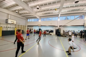 Mecz siatkówki rozgrywany w szkolnej sali gimnastycznej, gdzie zawodnicy w czerwonych i białych koszulkach skupieni są na piłce lecącej nad siatką, a na trybunach w tle zgromadzeni są kibice.