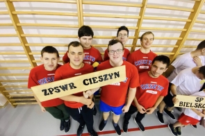 Grupa zawodników w czerwonych koszulkach sportowych stoi w sali gimnastycznej przy drabinkach, trzymając drewnianą tabliczkę z nazwą ZPSWR Cieszyn i uśmiechając się do zdjęcia.