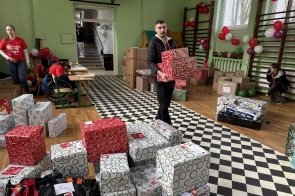 Mężczyzna niosący kilka paczek zapakowanych w świąteczny papier, przechodzący przez salę gimnastyczną, w której na podłodze i w tle ustawione są kolejne paczki oraz kartony, a przestrzeń ozdobiona jest biało-czerwonymi balonami.