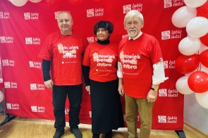 Po lewej stronie stoi Jarosław Bodak, Wiceprzewodniczący Rady, pośrodku Janina Żagan, Starosta Cieszyński, a po prawej Stanisław Kubicius, Przewodniczący Rady. Wszyscy ubrani są w czerwone koszulki z napisem „Człowiek pełen dobra”. W tle widoczna jest czerwona ścianka z hasłem „lubię ludzi” oraz logotypem Szlachetnej Paczki, otoczona biało-czerwonymi balonami.