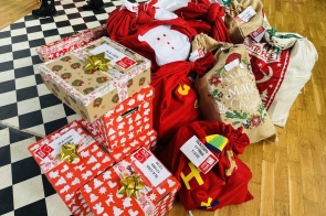 Paczki zapakowane w świąteczny papier oraz czerwone i jutowe worki z motywami bożonarodzeniowymi, ułożone na podłodze sali gimnastycznej z widoczną szachownicą i drewnianą podłogą.
