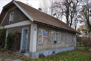 Domek Ogrodnika od strony ulicy/wejścia, niewielki zabytkowy budynek z jasną elewacją, spadzistym dachem i oknami zabezpieczonymi kratami, otoczony zielenią.