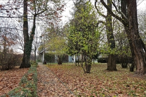 Fragment dawnego parku dworskiego, z tyłu widoczny Domek Ogrodnika, otoczony drzewami i jesiennymi liśćmi.