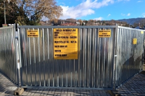 Metalowe ogrodzenie placu budowy z zamontowanymi żółtymi tablicami informacyjnymi i ostrzegawczymi. Na środku tablica budowy, po bokach napisy o zakazie wstępu i parkowania. W tle drzewa i zabudowania.