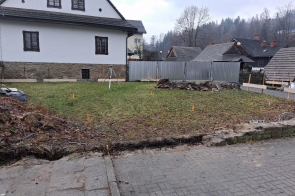 Teren zielony w trakcie prac ziemnych przed budynkiem. Widoczne paliki geodezyjne, statyw pomiarowy oraz fragment wykopu przy chodniku. W tle metalowe ogrodzenie i drewniane zabudowania.