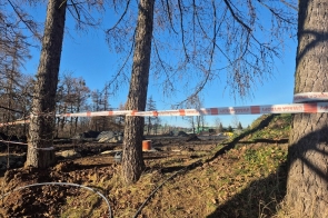 Teren robót ziemnych wśród drzew, ogrodzony taśmą ostrzegawczą, z widocznymi wykopami, rurami i pryzmami ziemi; w tle elementy infrastruktury, przy słonecznej pogodzie.