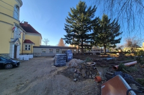 Dziedziniec przy budynku, na którym prowadzone są prace budowlane; widoczne są składowane materiały, gruz, rury oraz zaparkowany samochód, a całość otaczają drzewa przy bezchmurnym niebie.