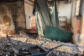 Pomieszczenie w trakcie prac budowlanych. Podłoga pokryta gruzem i fragmentami tynku. Na pierwszym planie taczka wypełniona odpadami budowlanymi. W tle ściana z odkrytymi instalacjami, częściowo zdemontowanymi elementami oraz zasłoniętym folią fragmentem pomieszczenia. Widoczne są odsłonięte cegły, rury oraz narzędzia opierające się o ścianę.