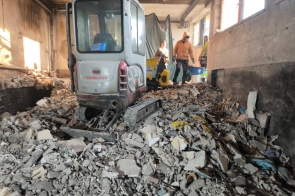 Wnętrze budynku w trakcie prac rozbiórkowych. Na pierwszym planie znajduje się mała koparka gąsienicowa stojąca na gruzie budowlanym złożonym z fragmentów betonu, tynku i innych materiałów. W tle widoczne są trzy osoby wykonujące prace porządkowe, wyposażone w odzież roboczą oraz narzędzia, w tym taczki. Pomieszczenie ma nieukończone ściany i sufit, a światło wpada przez okna po prawej stronie.