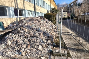 Na pierwszym planie znajduje się duża hałda gruzu budowlanego złożona z fragmentów betonu, cegieł i innych materiałów, usypana przy ścianie budynku. Po prawej stronie widoczne jest metalowe ogrodzenie tymczasowe oddzielające teren robót od chodnika i drogi. W tle znajduje się kilkukondygnacyjny budynek w jasnych kolorach z rzędami okien oraz drzewa bez liści. Zdjęcie wykonane w słoneczny dzień.