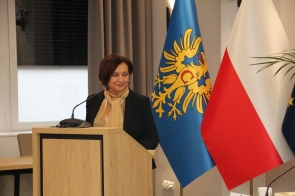 Janina Żagan Starosta Cieszynski przemawia; ubrana w ciemną marynarkę i jasny szal. W tle widoczne są flagi oraz zasłona.