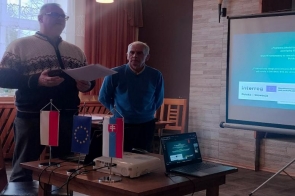 Dwóch mężczyzn stoi obok siebie, jeden z nich trzyma dokumenty. Na stole znajduje się projektor, laptop oraz małe flagi Polski, Unii Europejskiej i Słowacji. Obok ustawiony jest ekran z wyświetloną prezentacją programu Interreg Polska-Słowacja. W tle widoczne są krzesła, okno z zasłonami i oświetlenie sufitowe.