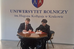 Dwie osoby siedzące przy ozdobnym, ciemnym stole i podpisujące dokumenty w czerwonych teczkach. W tle jasna ściana z herbem oraz napisem „Uniwersytet Rolniczy im. Hugona Kołłątaja w Krakowie”. Osoby ubrane są w stroje formalne, na stole leżą dokumenty i długopisy.