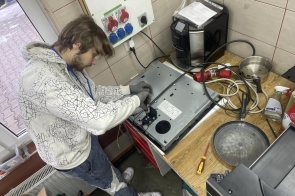 Młody mężczyzna w jasnej bluzie i rękawicach roboczych pracuje przy rozebranym urządzeniu elektronicznym leżącym na drewnianym blacie. Skupiony pochyla się nad wnętrzem sprzętu, przy którym widać przewody i podzespoły. W tle znajduje się ściana z gniazdami przemysłowymi, a na stole leżą kable, wtyczki oraz narzędzia.