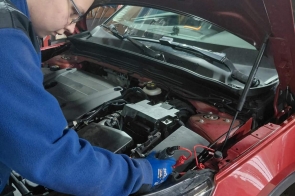 Młody mężczyzna w niebieskiej bluzie i okularach pochyla się nad otwartą maską czerwonego samochodu, pracując przy elementach silnika. Jedną ręką sięga do komory silnika, w której widoczne są akumulator, przewody i metalowe podzespoły.