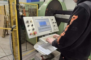  Młody mężczyzna w ciemnej bluzie i spodniach roboczych stoi przy dużym urządzeniu technicznym i obsługuje panel sterowania z przyciskami oraz pokrętłami. Obok znajduje się metalowa osłona z siatki, a w tle widać przewody i elementy konstrukcyjne pomieszczenia.