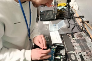 Młody mężczyzna w jasnej bluzie z identyfikatorem na niebieskiej smyczy pochyla się nad rozebranym laptopem na stole. Skupiony pracuje przy płycie głównej i podzespołach urządzenia, a w tle widać drugi laptop, przewody oraz sprzęt elektroniczny.