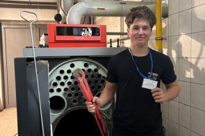 Młody mężczyzna w czarnym stroju roboczym stoi w kotłowni obok dużego kotła grzewczego z otwartą komorą. Ma na szyi identyfikator na niebieskiej smyczy i trzyma w ręku czerwony klucz nastawny. W tle widać białe rury instalacyjne oraz ściany wyłożone jasnymi płytkami.