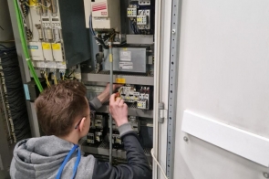 Młody mężczyzna w szarej bluzie z kapturem i identyfikatorem na niebieskiej smyczy pracuje przy otwartej szafie sterowniczej. Z tyłu, z profilu, reguluje lub podłącza elementy instalacji elektrycznej, w której widać moduły, przekaźniki, przewody oraz urządzenia automatyki.