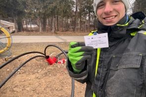 Młody mężczyzna w szarej czapce, ciemnej kurtce roboczej z elementami odblaskowymi oraz zielono-czarnych rękawicach trzyma w dłoni identyfikator. Obok niego widoczny jest przewód zakończony czerwoną wtyczką przemysłową. W tle widać drogę oraz drzewa bez liści.