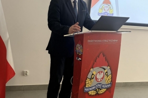 Mieczysław Szczurek Wicestarosta Cieszyński w garniturze i krawacie stoi na czerwonym podium z napisem "Państwowa Straż Pożarna" oraz emblematem formacji, mając przed sobą otwarty laptop. W tle ekran prezentacyjny z elementem graficznym w barwach biało-czerownych.