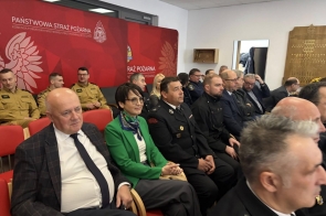 Rząd osób siedziących na czerwonych krzesłach, wśród nich mężczyźni w mundurach strażakich. W tle czerowna ścianka. W sali widoczne jasne ściany i sufit z panelowym oświetleniem.