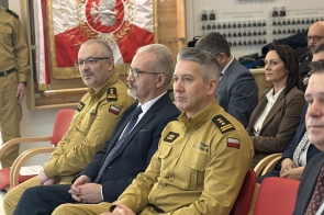 Pierwszy rząd uczestników siedziących na czerwonych krzesłach, wśród nich dwóch mężczyzn w żółtych mundurach strażackich oraz mężczyzna w garniturze i krawacie. W tle fragment czerwonego sztandaru z białym orłem oraz kolejne osoby w strojach mundurowych i eleganckich.