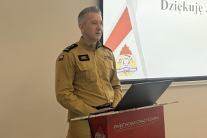  Roman Klecha Zastępca Śląskiego Komendanta Wojewódzkiego PSP w żółtym mundurze stoi przy czerwonym podium. W tle ekran prezentacyjny.