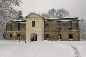 Budynek z centralną jasną częścią i przejazdem w formie łuku, otoczony śniegiem. Na dachu i parapetach widoczne są warstwy śniegu, a w tle znajdują się drzewa bez liści. Przed budynkiem widać zasypaną ścieżkę oraz znaki przy ogrodzeniu.