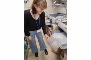 Kobieta stoi przy stole i przegląda materiały. Na blacie znajdują się kartki papieru i inne przedmioty. W tle widoczne są krzesła oraz wyposażenie pomieszczenia.