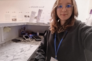 Kobieta stoi w pomieszczeniu biurowym, ma rozpuszczone włosy i okulary. Ma na szyi identyfikator. W tle widoczne są dokumenty oraz sprzęt biurowy ustawiony na blatach i półkach.