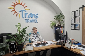 Mężczyzna siedzi przy biurku w biurze. Przed nim znajduje się komputer, dokumenty i telefon. Na ścianie widoczne jest logo firmy, a w pomieszczeniu stoją rośliny doniczkowe oraz sprzęt biurowy.