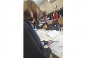 Kobieta siedzi przy stole i przegląda dokumenty. Na blacie leżą kartki papieru oraz przybory do pisania. W tle widoczne są krzesła i elementy wyposażenia pomieszczenia.