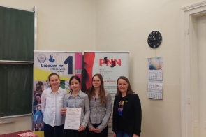 Jasna sala z wysokimi ścianami, tablicą po lewej i drzwiami po prawej. Na ścianie wisi czarny zegar i kartkowy kalendarz. Cztery kobiety stoją obok siebie na parkiecie, przed dwoma stojącymi banerami: „Liceum nr 1 w Cieszynie” oraz „PSNn”. Jedna z nich trzyma w rękach dyplom.