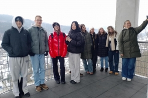 Taras widokowy z metalową barierką i betonowym słupem. Grupa młodych osób w zimowych kurtkach stoi obok siebie i patrzy w stronę aparatu; jedna z nich po prawej stronie trzyma wyciągnięty telefon. W tle widoczny jest zimowy krajobraz z drzewami i zabudowaniami w dolinie.