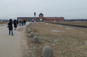 Szeroka droga prowadząca w stronę ceglanych budynków. Po lewej stronie idzie grupa osób w zimowych kurtkach. Po prawej widać tory kolejowe biegnące równolegle do drogi oraz rozległy, pusty teren z łatami śniegu. Wzdłuż drogi ustawione są kamienne słupki.