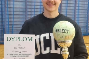 Młody mężczyzna stoi na środku kadru i uśmiecha się do aparatu. W jednej ręce trzyma dyplom z napisem „DYPLOM”, a w drugiej wysoki puchar zakończony piłką.