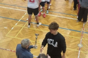 Na pierwszym planie starszy mężczyzna wręcza młodemu mężczyźnie puchar, obok stoi druga osoba trzymająca dyplom. W tle ustawiona jest grupa chłopców w białych koszulkach, a część osób stoi z boku i obserwuje wydarzenie.