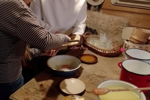 Drewniane wnętrze z rustykalnym stołem pełnym naczyń i produktów. Dwie młode dziewczyny stoją obok siebie i kroją jasny blok sera na drewnianej desce. Na stole widać m.in. emaliowane garnki, miskę, talerzyki, koszyk ze szklankami oraz kawałek sera na spodku. W tle znajdują się drewniane ściany, lustro i wiszący kapelusz.