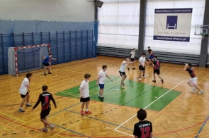 Hala sportowa z drewnianą podłogą i bramką do piłki ręcznej po lewej stronie. Na boisku trwa mecz; zawodnicy w białych koszulkach bronią, a zawodnicy w czarno-czerwonych koszulkach rozgrywają akcję w ataku. Bramkarz stoi przed bramką z uniesionymi rękami, a po prawej stronie widoczny jest sędzia w żółtej koszulce. W tle znajdują się trybuny oraz baner reklamowy.