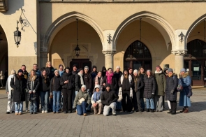 Jasny plac przed zabytkowym budynkiem z arkadami i ozdobnymi lampami. Duża grupa osób w zimowych kurtkach, czapkach i szalikach stoi w kilku rzędach i pozuje do wspólnego zdjęcia. W tle widać kamienne kolumny, wejścia do budynku oraz dekoracyjne detale architektoniczne.