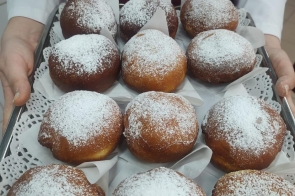 Metalowa taca wypełniona dwunastoma pączkami posypanymi cukrem pudrem, ułożonymi na białych serwetkach. Tacę trzyma osoba ubrana w białą koszulę i czerwony fartuch. Pączki mają złocistobrązową skórkę i są równomiernie oprószone cukrem.