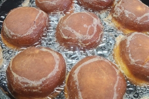 Osiem pączków smażących się w głębokim oleju na dużej patelni. Ciasto ma złocistobrązową skórkę z jasną obwódką pośrodku. Wokół pączków intensywnie bulgocze olej. W tle widoczny fragment czerwonego fartucha i białego rękawa.