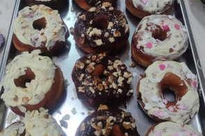 Metalowa taca z kilkunastoma pączkami z otworem w środku. Część oblana ciemną polewą czekoladową i posypana kawałkami orzechów, część pokryta jasną polewą z płatkami migdałów lub różowo-białą posypką w kształcie serduszek. Pączki ułożone w równych rzędach.