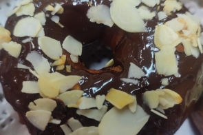 Pączek z otworem w środku oblany błyszczącą polewą czekoladową i posypany płatkami migdałów. Leży na metalowej tacy wyłożonej białą serwetką. Na dole kadru widoczny fragment innego pączka oprószonego cukrem pudrem.