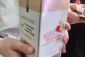 Dwie osoby trzymają tekturową puszkę kwestarską Fundacji „Serca dla Maluszka”. Jedna z nich wkłada zwinięty banknot do otworu w górnej części puszki. Na obudowie widoczny jest numer puszki, nazwa akcji „Cała Polska smaży pączki 2026” oraz logo fundacji.