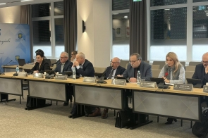 Kilku radnych siedzi przy długim stole konferencyjnym w sali sesyjnej. Przed każdą osobą stoją tabliczki z imionami i nazwiskami oraz mikrofony, na blacie znajdują się także butelki z wodą i dokumenty. W tle widoczne są duże okna z roletami, zasłony oraz godła na ścianie i baner z herbem.