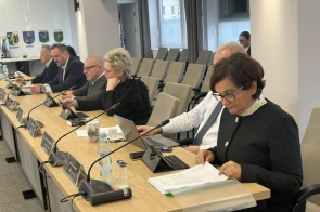 Kilku radnych siedzi przy długim stole w sali sesyjnej i przegląda dokumenty. Po prawej stronie widoczna jest m.in. Janina Żagan Starosta Cieszyński ubrana w czarną marynarkę, obok niej Mieczysław Szczurek Wicestarosta Cieszyński w białej koszuli i krawacie, dalej Beata Macura Członkini Zarządu w czarnej bluzce oraz Marcin Ślęk Członek Zarządu w szarym swetrze. W dalszej części stołu siedzą kolejni radni.Na blacie znajdują się mikrofony i tabliczki z imionami i nazwiskami. W dalszej części stołu siedzą kolejni radni. 